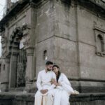 A couple in traditional white clothing sitting by a historic stone building outdoors.